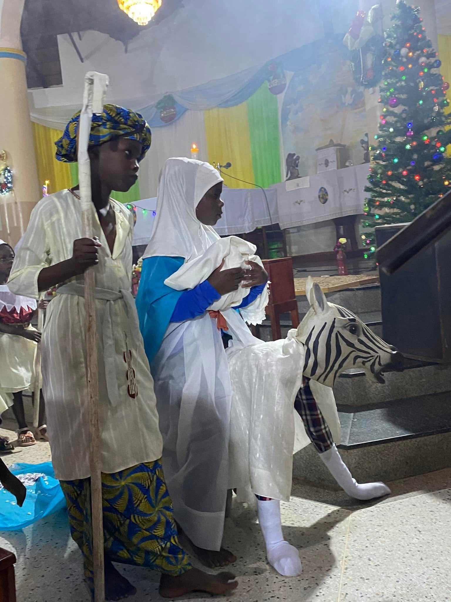 Mercy School children perform a nativity play in a church: a shepherd, Mary holding a baby, and a child in a zebra costume