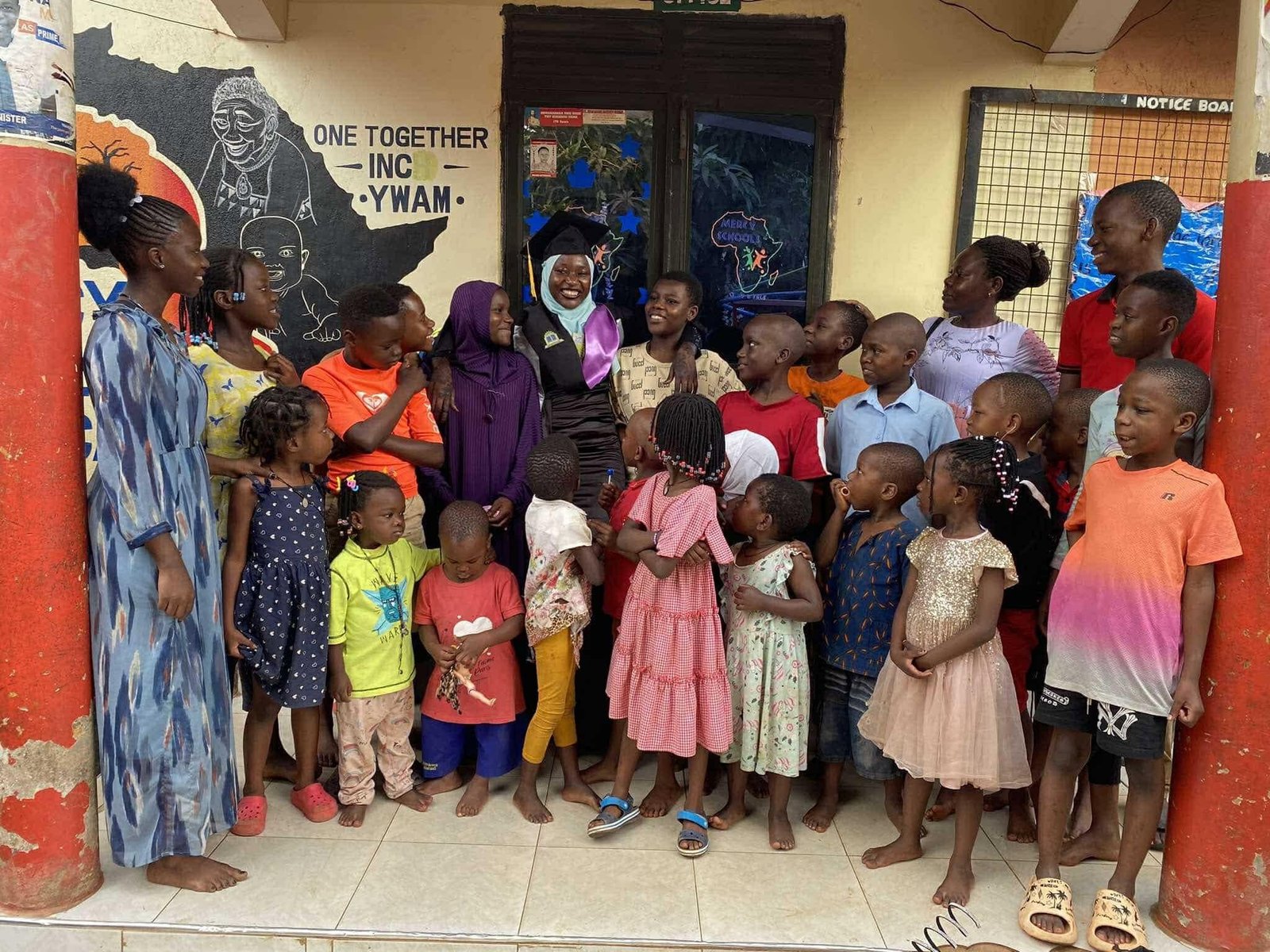 A graduate in a cap and gown stands on the Mercy School porch surrounded by smiling children